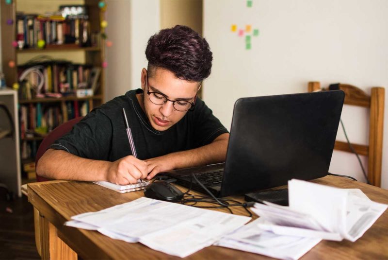 A young man writing in a notebook while seated at a table topped with paperwork and a laptop