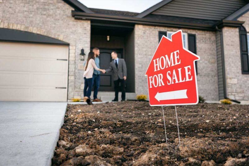 A young couple shake hands with a real estate agent in front of a home for sale.